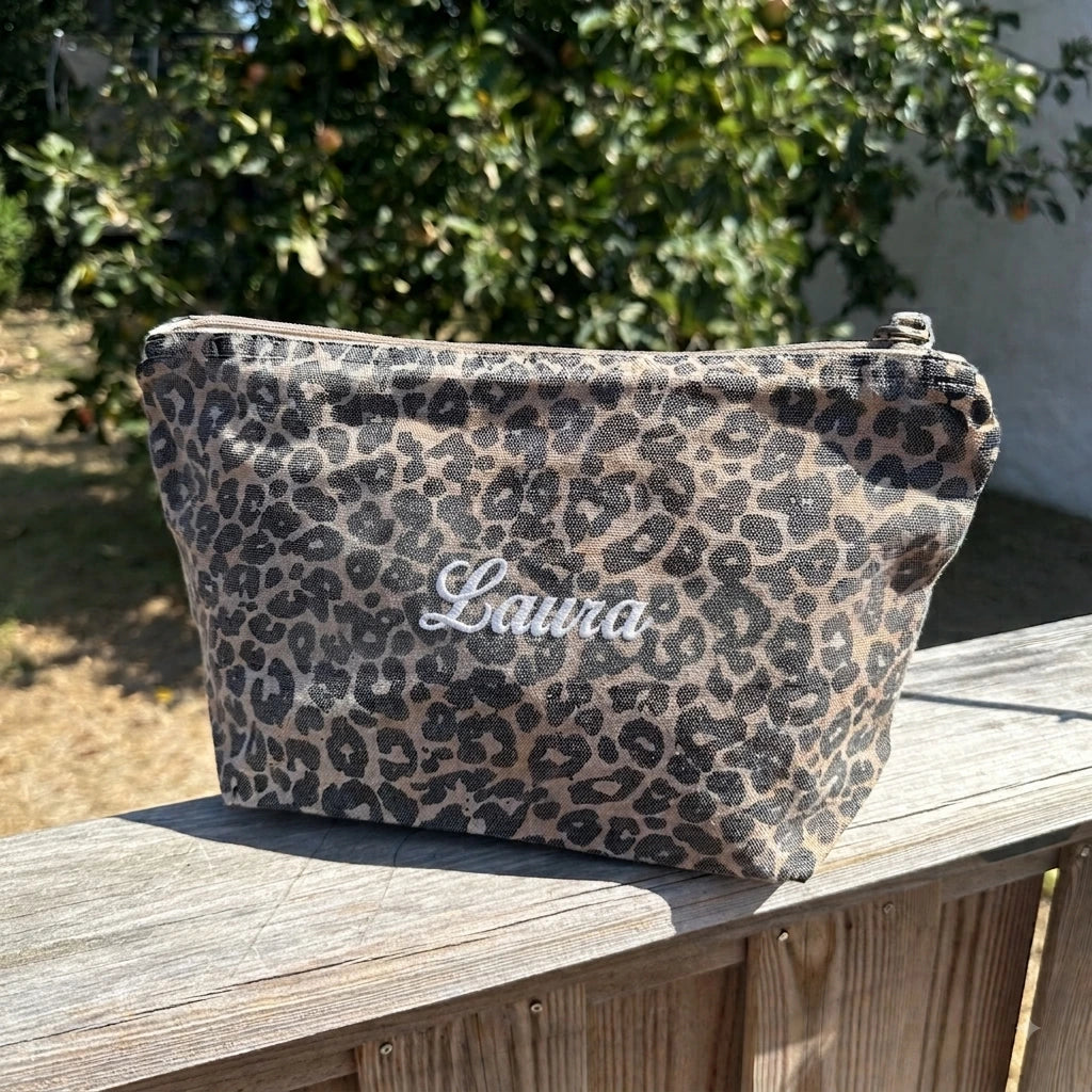 A leopard-print cosmetic pouch with the name 'Laura' embroidered in white script, resting on a wooden deck railing with a garden background.