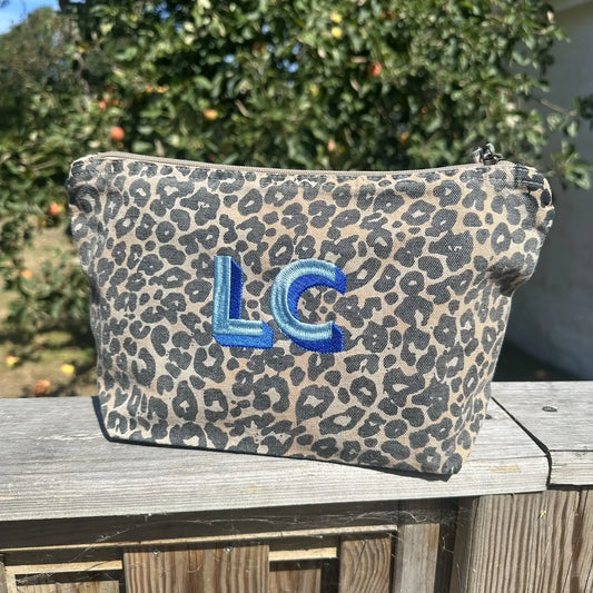 A leopard print cosmetic pouch with a blue embroidered monogram 'LC' sits on a wooden deck railing with an outdoor garden background.